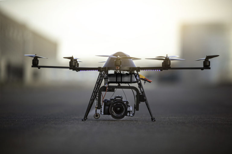 Bild: Drohne Multicopter mit Kamera, zeigt eine Drohne mit mehreren Rotoren am Boden stehend und eine Kamera im Schlepptau.